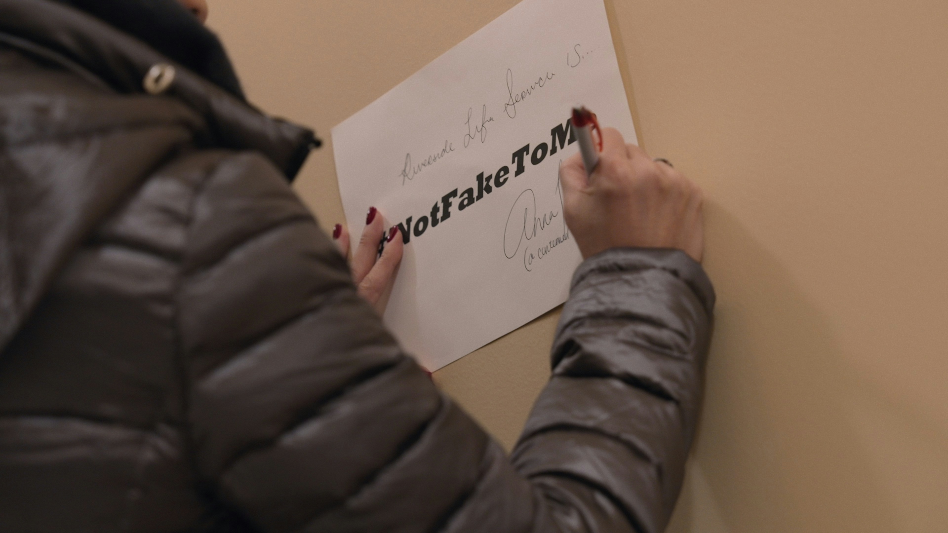 A woman signing a piece of paper with "Not Fake To Me" written on it